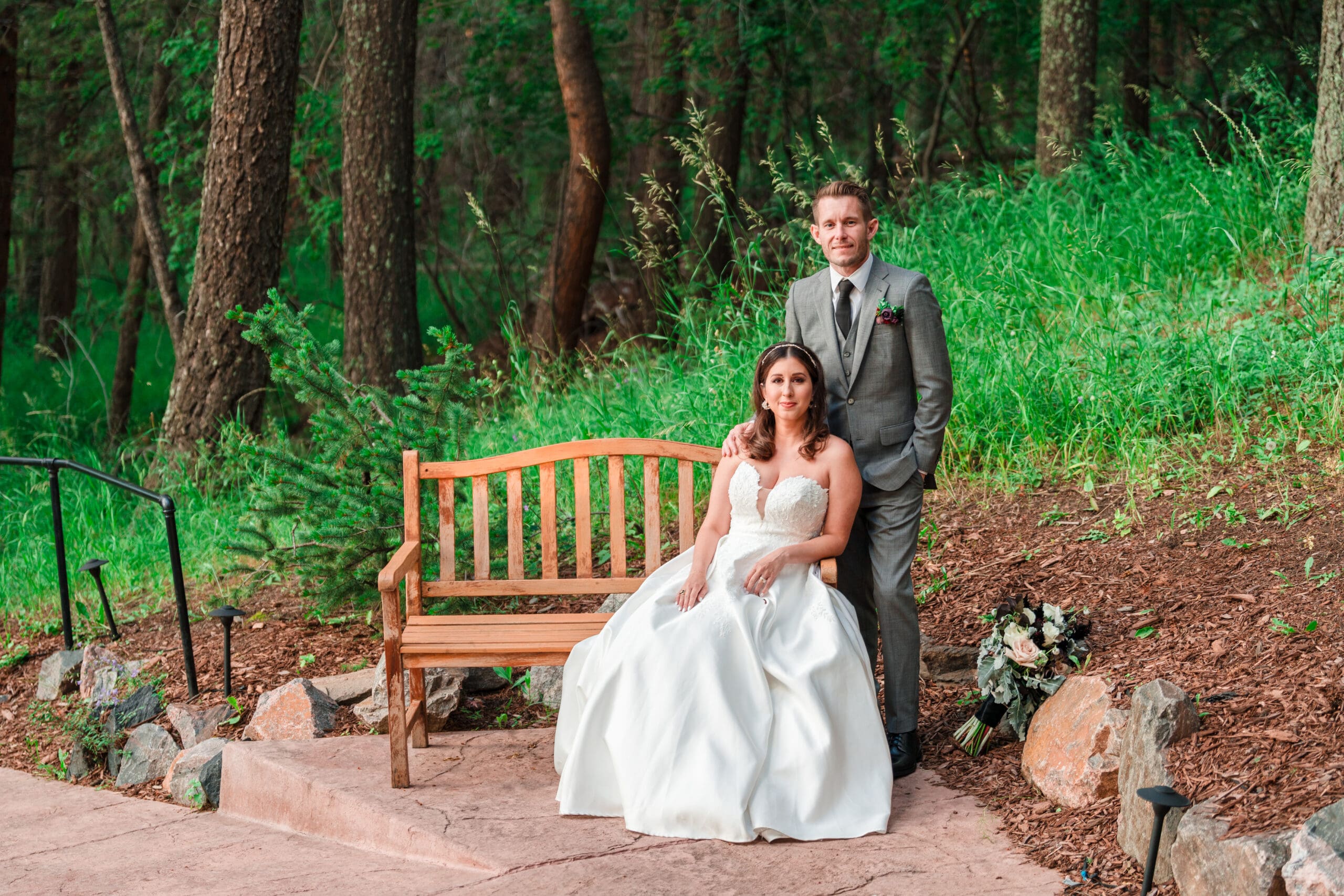Destination wedding bliss: Bride sits on a bench with her husband standing beside her, symbolizing their love and unity on their special day.