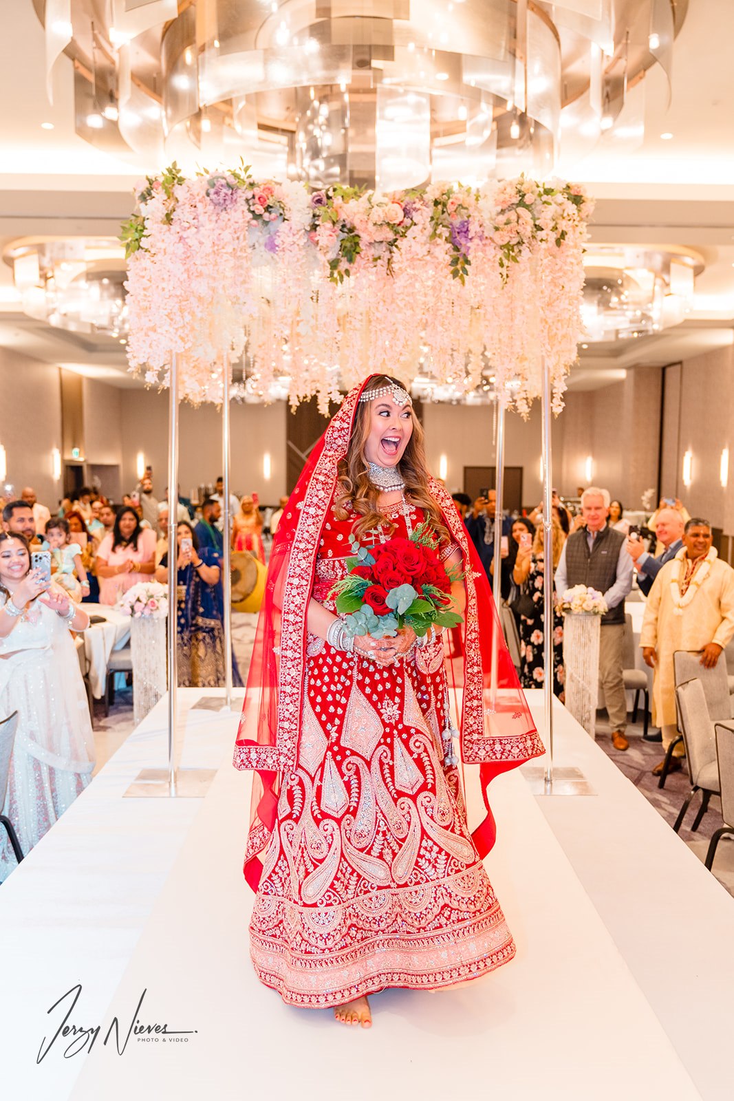Nicole walking down the runway adorned in a traditional red Indian wedding dress, holding red flowers, as guests stand in reverence at Disney Swan Reserve.