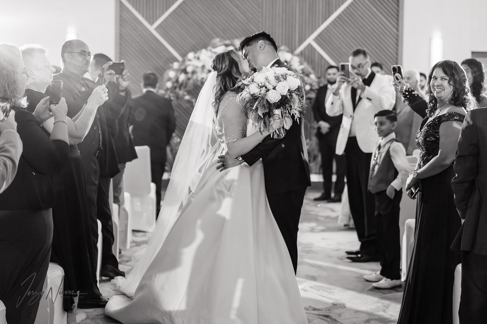 Black and white photo of Ravin and Nicole sharing a kiss at the Disney Swan Reserve ceremony, walking down the aisle as husband and wife, while guests smile and take pictures.