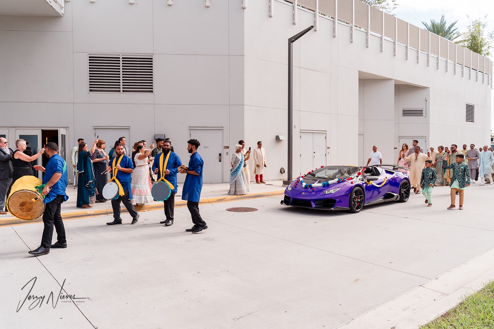 Wide shot of the parade procession with the band leading the Lamborghini and the wedding party walking behind it.