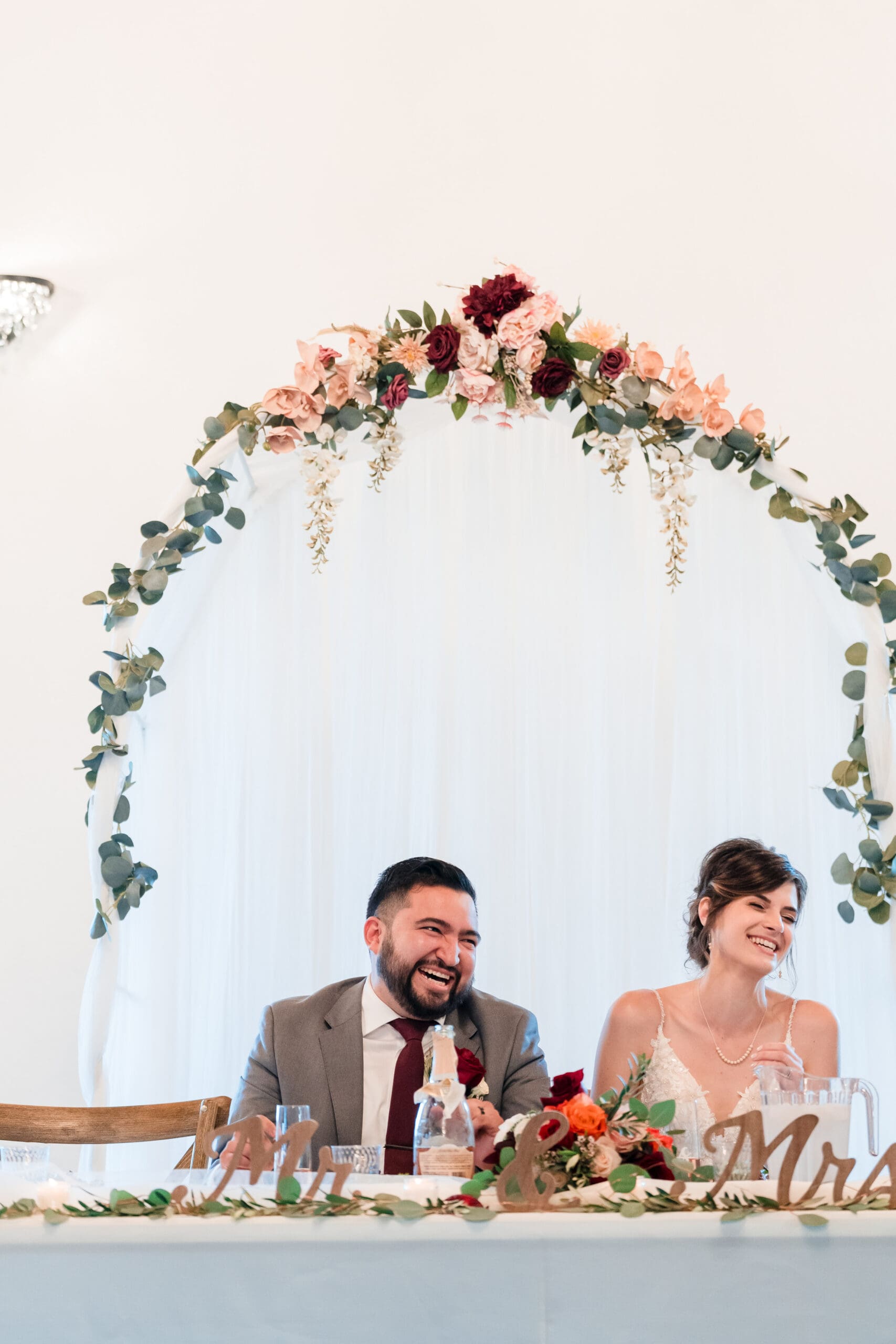 A tall shot capturing the newlyweds seated at the table on the stage, adorned with "Mr & Mrs" decorations, both beaming with joy during the reception at the Sterling Event Venue Reception Center.