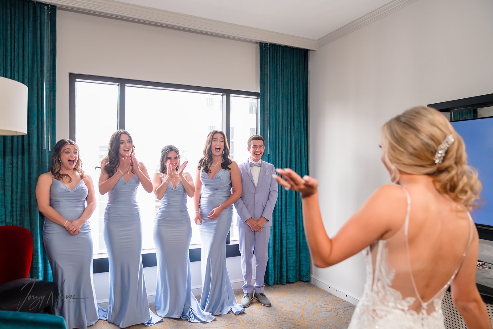 Bridal party's emotional reaction to the bride in her dress after hair and makeup, captured from behind the bride with a focus on their surprised and joyful expressions.