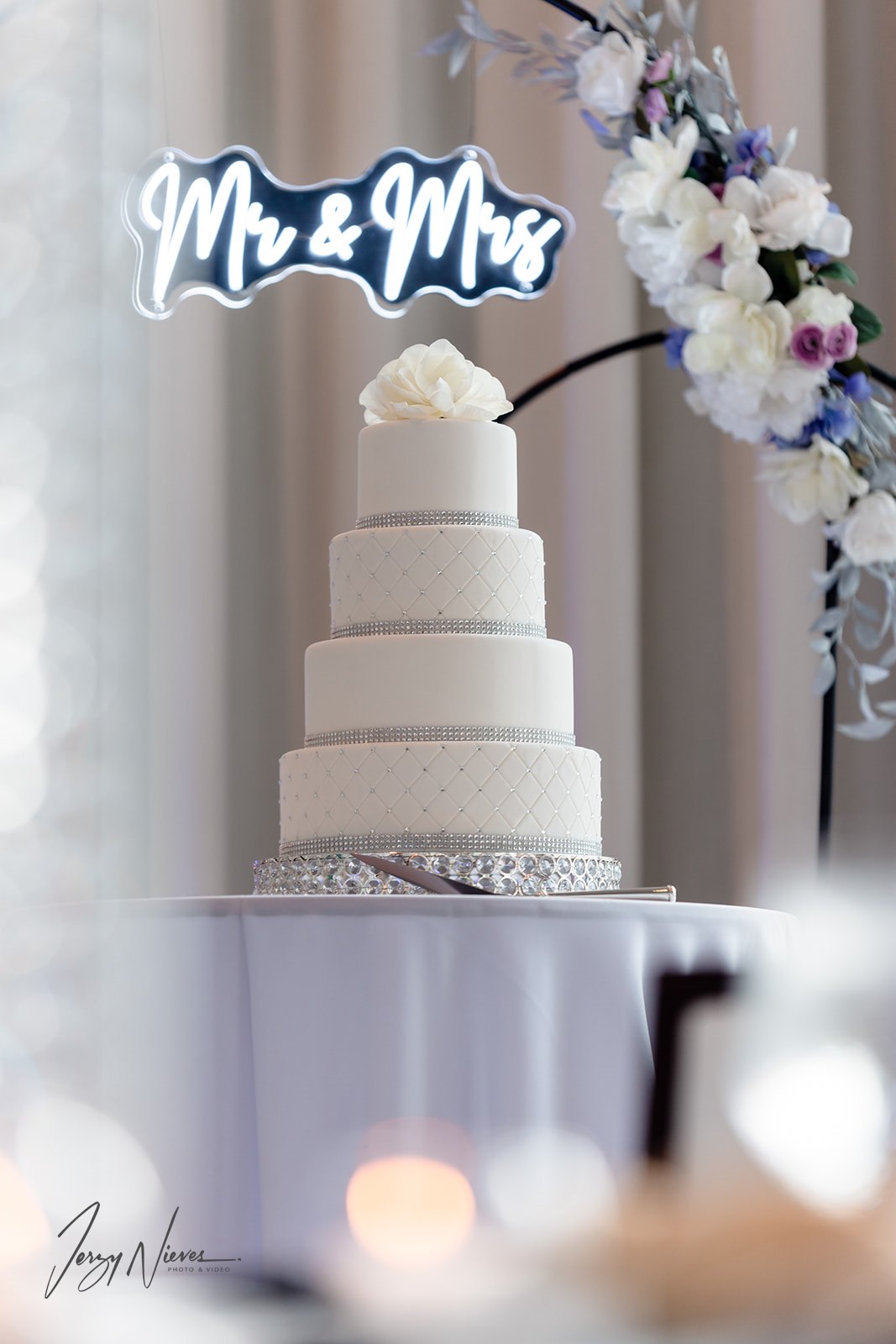 Close-up of a four-tier wedding cake with a "MR & MRS" light illuminating in the background.