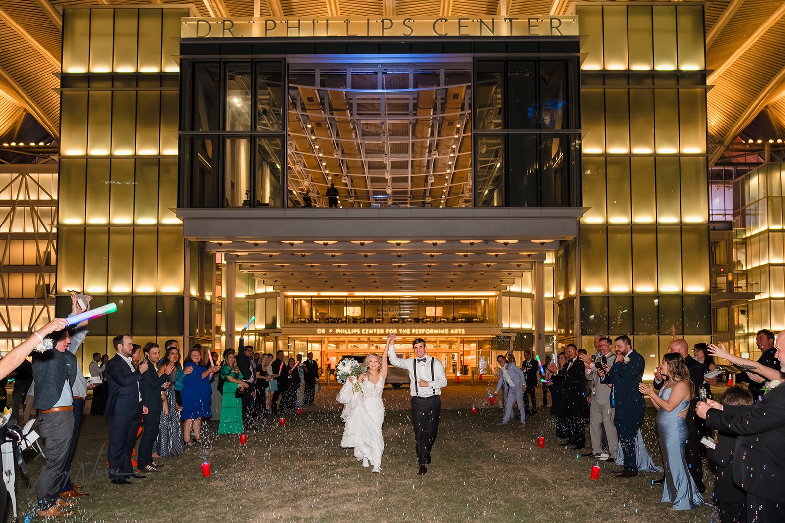 Colorful image of the bride and groom walking down the grand exit, holding hands with hands raised between them, surrounded by bubbles and guests clapping in celebration in front of the Dr Phillips Performing Arts Center.