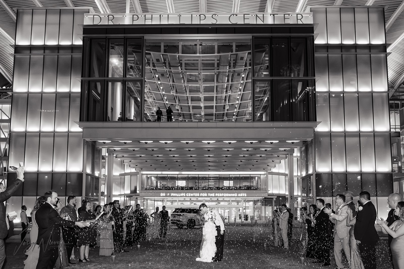 Black and white image of the bride and groom kissing in front of the Dr Phillips Performing Arts Center during their grand exit, surrounded by guests blowing bubbles.