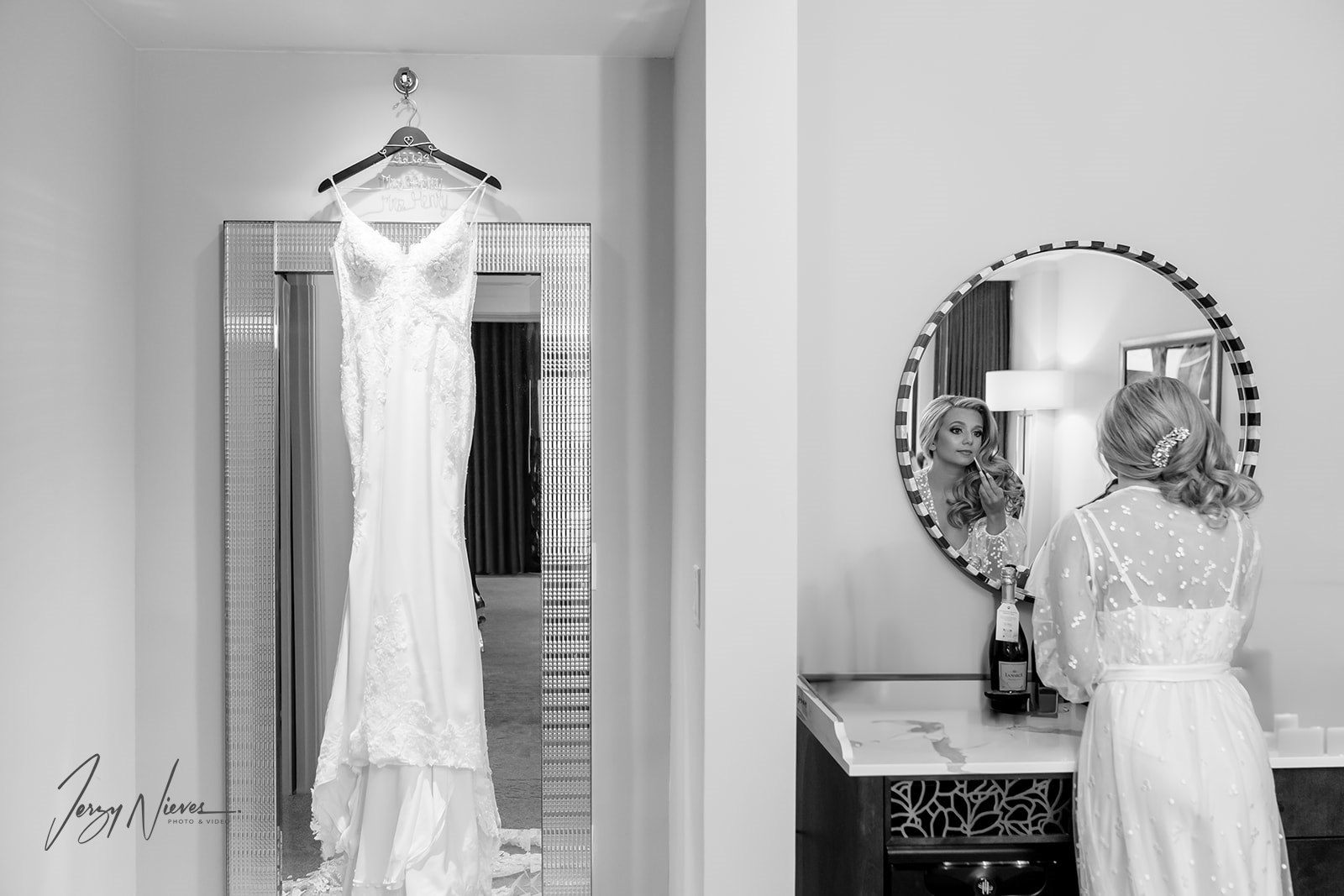 Black and white image of the bride finishing her preparations in front of a mirror, with her wedding dress hanging beside her.