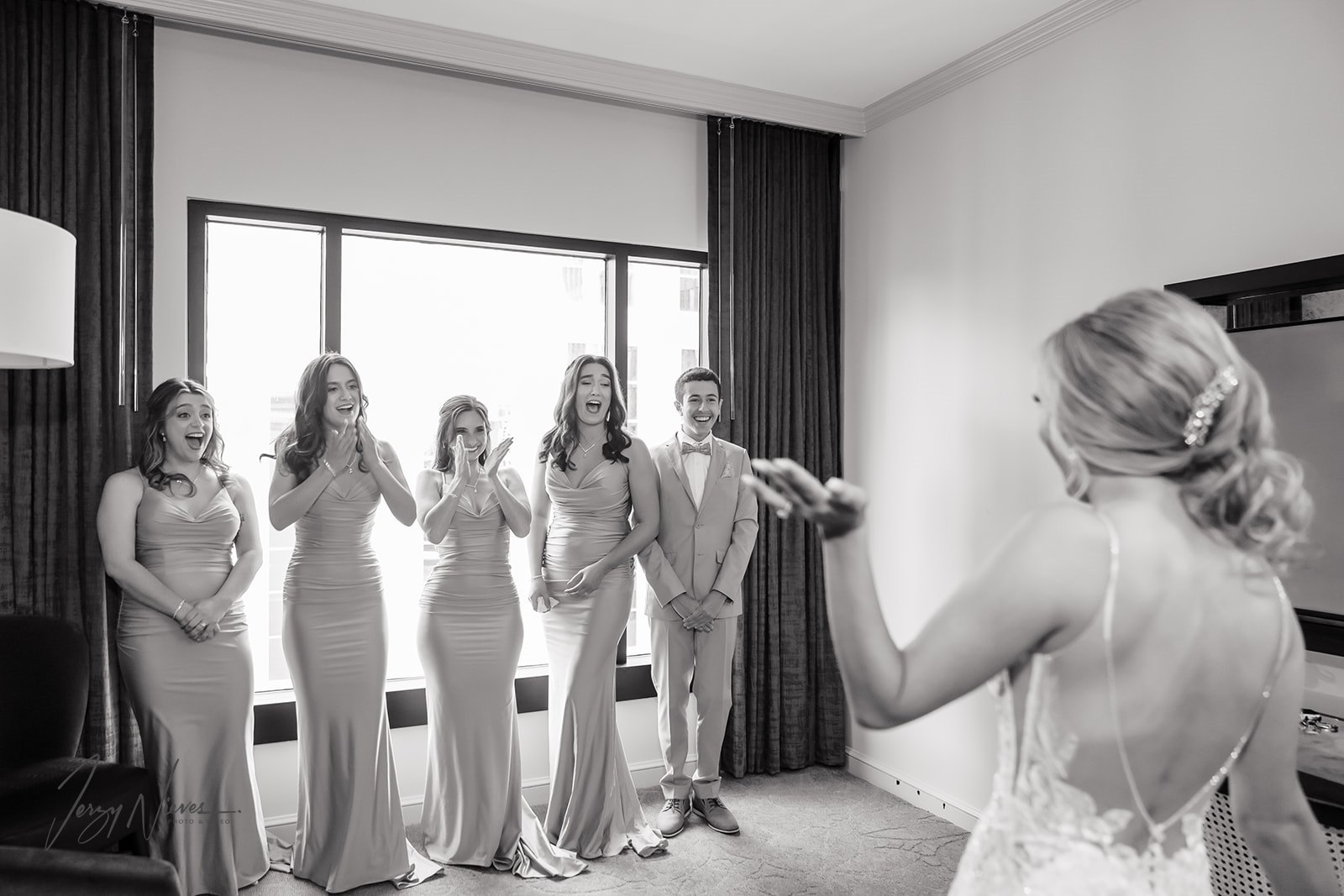 Black and white photograph capturing the emotional reaction of the bridal party as they see the bride in her dress after hair and makeup, taken from behind the bride.